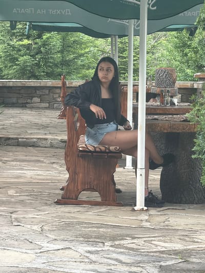 A young woman is captured sitting relaxed on a rustic wooden bench in an outdoor setting. She is positioned towards the left of the frame, looking directly at the camera with a neutral expression. She has dark hair, wears a long-sleeved dark top, and light blue denim shorts. Her legs are crossed, with one foot resting on a white horizontal bar. A small, dark tattoo is visible on her upper right thigh. She is wearing black sneakers.

The setting appears to be an outdoor café or restaurant area, possibly in a garden or courtyard. The ground is paved with light-colored, irregular stone slabs. In the background, lush green trees and foliage are visible, suggesting a natural environment. To her right, a rustic wooden table with carved details holds several glass bottles. A large green umbrella, partially visible on the left, provides shade. The umbrella features inverted and reflected text in Cyrillic script, likely part of a brand name or establishment name. White support poles extend vertically in front of the woman. The scene is brightly lit, indicating it is daytime, likely late morning or afternoon with clear weather. The overall ambiance is relaxed and natural.