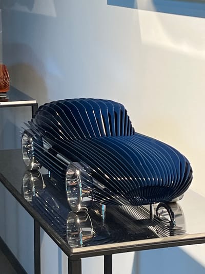 The image shows a contemporary art sculpture of a car, made of stacked, curved dark blue layers. The sculpture is placed on a glossy black surface, likely a display table, which reflects the object. The car has shiny, metallic wheels and sits on thin chrome legs. In the background, a white wall is visible with soft lighting and shadows, suggesting an indoor exhibition space. A hint of another object, possibly a decorative vase with an orange ribbed pattern, can be seen on a shelf to the left. The setting appears to be a modern gallery or showroom. The location is identified as Reckange-sur-Mess, Luxembourg.
