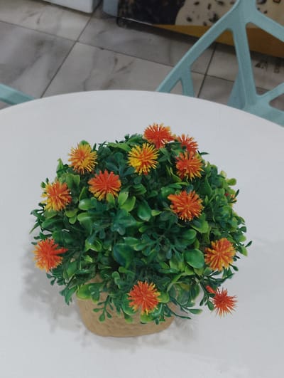 A close-up, overhead shot captures a small, artificial potted plant with vibrant orange and yellow flowers and green leaves. The plant sits on a white circular surface, possibly a table, in what appears to be an indoor setting. In the background, a light blue chair with a distinctive crisscross design is partially visible. The floor is tiled with light gray and white square tiles. The overall lighting suggests it is daytime. This scene is located in El Espinal, Venezuela.