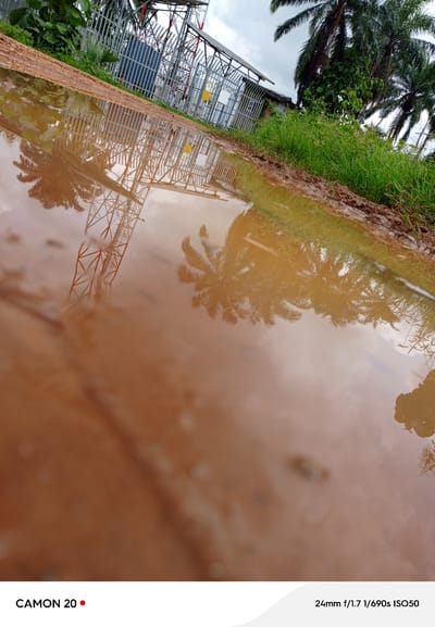 The image shows a muddy puddle reflecting the sky and a metal tower structure in Kaninkon, Nigeria. Palm trees and green vegetation are visible in the background. The puddle is a reddish-brown color, suggesting it's a mixture of mud and water. The metal tower appears to be part of a telecommunications or power transmission facility, with intricate lattice work. The sky is overcast, casting a diffused light on the scene. The overall impression is of a rural or semi-urban industrial area after rain.