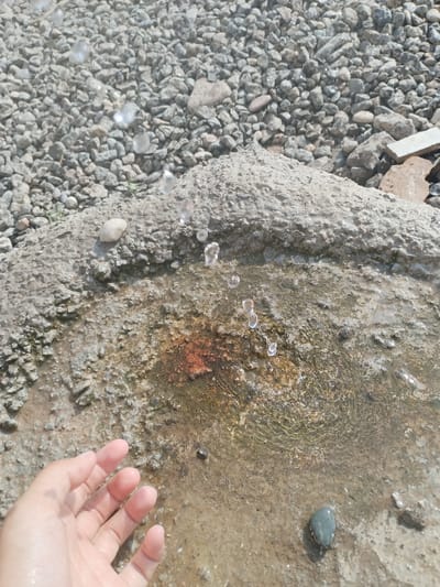 A person's left hand is visible in the lower left corner of the frame, reaching towards a puddle of water. The hand is pale and appears wet, with fingers slightly splayed. The puddle is located on a rough, textured surface that seems to be a mix of concrete and dirt, with a reddish-brown stain in its center. Several water droplets are suspended in the air above the puddle, indicating that water is actively falling or splashing. The background consists of a bed of small, grey pebbles and some larger rocks. The lighting suggests it is daytime and sunny. The scene is likely an outdoor area, possibly a street or public space, in Mady, Kyrgyzstan.