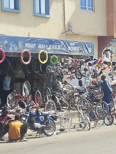 This image depicts a street scene in Darmanawa, Nigeria, centered on an outdoor repair and sales establishment for bicycles and motorcycles. The business operates under a blue awning displaying the name "YUSUF MAI-ZARE & SONS," affixed to a light-colored, two-story building featuring blue-framed windows on its upper floor.
The shop's exterior is densely populated with merchandise. Numerous bicycle wheels, tires, and frames are visible, some stacked, others hanging, and integrated into partially assembled or complete bicycles, including smaller models. Decorative tire casings in red, yellow, and green hang from the awning structure. Various smaller bicycle components, such as handlebars and seats, are mounted on display racks.
On the street level in the foreground, a dark-colored motorcycle, featuring blue and red accents, is parked on its kickstand. Two adult males are squatting beside it; one wears a red shirt, the other an orange shirt, both with dark trousers. Further back within the shop's display area, a third adult male in a light-colored shirt is seated. To the right of the scene, a fourth adult male, dressed in a dark blue tunic, stands facing towards the shop's interior displays.