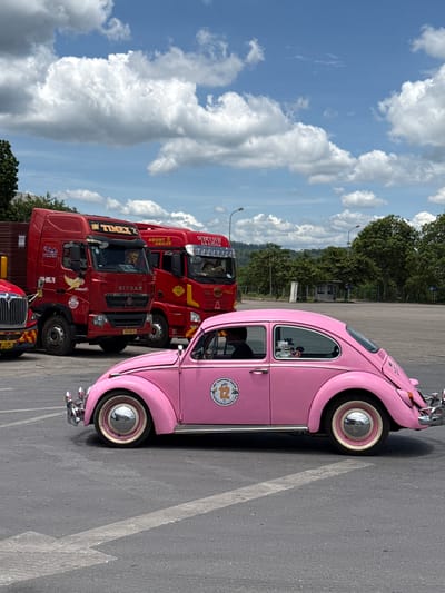 A pink Volkswagen Beetle is parked next to several red trucks. The Beetle is classic and has chrome accents and whitewall tires. The trucks are large commercial vehicles with various logos and text visible. The scene appears to be a parking lot or an area with asphalt road markings. In the background, there are trees and a cloudy sky, suggesting it is daytime. The image is oriented vertically, as if taken by someone holding their phone in a portrait position. Text visible on the trucks includes "TIMEX", "SITRAK", and "KNIGHT HAULER". There are also license plate numbers like "15H-085.76". The overall atmosphere seems to be a casual outdoor scene, possibly a roadside stop or a parking area for freight vehicles.