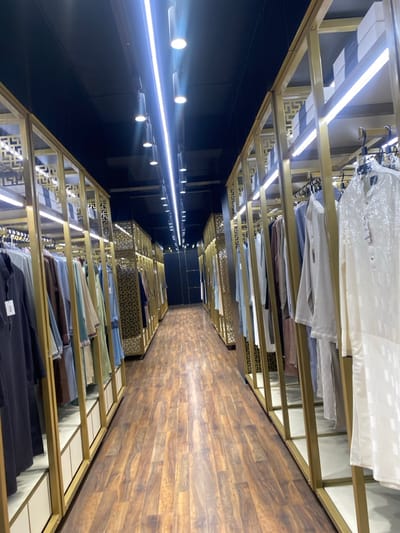 The image displays an interior view of a retail clothing store located in Narayanganj, Bangladesh. A long, narrow aisle extends from the foreground into the background, flanked by parallel clothing display units on both sides. The floor features dark brown, wood-patterned laminate. The display units consist of gold-colored metal frames with intricate geometric lattice designs, supporting shelves and garment rails.

Numerous traditional garments, likely kurtas or similar South Asian attire, are hung on hangers along both sides of the aisle. The clothing items exhibit a variety of colors including white, cream, light blue, green, beige, brown, and black. Some garments are protected by clear plastic covers, and visible tags are attached. Upper shelves on the display units hold stacks of white or light-colored rectangular boxes.

The ceiling is dark, possibly black, and illuminated by a prominent central linear LED light fixture running the length of the aisle. Additional black cylindrical spotlights are suspended from the ceiling, providing further illumination. The perspective is centered down the aisle, converging towards a darker area at the far end. No individuals are visible in the scene, and no specific actions or interactions are depicted.