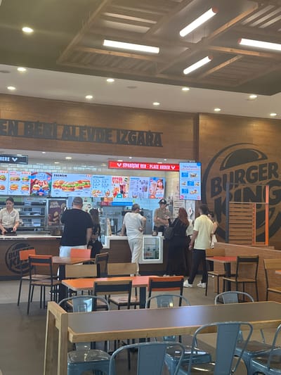 The image shows the interior of a Burger King restaurant, likely located in Manisa, Turkey, as the Turkish language is prominent. A line of people is visible, waiting to place orders at the counter, which features digital menu boards displaying food options. The restaurant design includes wood paneling, Burger King branding, and modern lighting. Seating is available at high tables with blue chairs. People appear to be engaged in the act of ordering food.