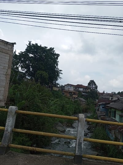 The image displays a residential area in Vieng Phouka, Laos, viewed from an elevated position, likely a bridge overlooking a ravine or riverbed. In the foreground, a concrete bridge railing with three horizontal yellow bars and grey vertical posts extends across the lower portion of the frame. Below the railing, a narrow, rocky stream flows through a vegetated channel. The water exhibits ripples, indicating current, and numerous stones are visible in the streambed.
Both banks of the stream are densely covered with lush green vegetation, including trees, bushes, and broad-leaved plants. On the left side of the scene, a partial view of an unfinished concrete structure with exposed blocks is visible. The background comprises a hillside densely packed with an assortment of residential buildings. These structures vary in design and condition, featuring a mix of painted walls, corrugated metal roofs, and terracotta-tiled roofs. Several prominent trees with full green canopies are interspersed among the dwellings. Multiple parallel black utility lines traverse the upper portion of the sky, which is overcast with grey and white clouds. No individuals are present in the scene.