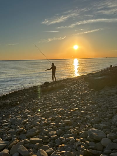 A person is fishing on a rocky beach at sunset in Blagoveshchenskaya, Russia. The sun is low on the horizon, casting a warm orange glow across the scene. The person, silhouetted against the bright sky, holds a fishing rod and appears to be casting a line into the calm sea. The beach is covered in smooth, gray pebbles. In the distance, several boats are visible on the water. The sky is a gradient of blue and orange, with wispy clouds scattered across it. The overall impression is one of tranquility and the beauty of nature.