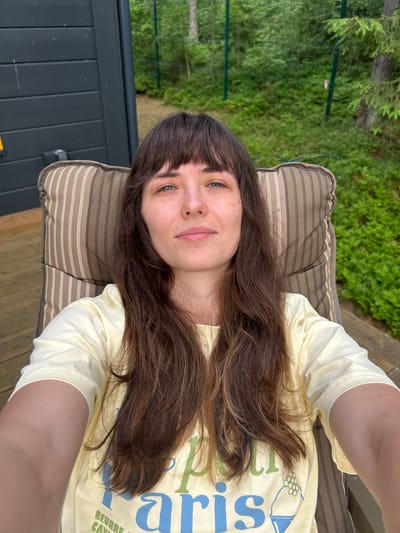 A woman with long brown hair and bangs sits in a striped outdoor chair on a wooden deck. She wears a pale yellow t-shirt with blue and green lettering. Behind her, a dark wooden wall of a building is visible on the left, and lush green foliage, including trees and bushes, fills the background. A green fence is also present in the background. The image appears to be a selfie taken outdoors. The location is stated to be Shepelyovo, Russia.