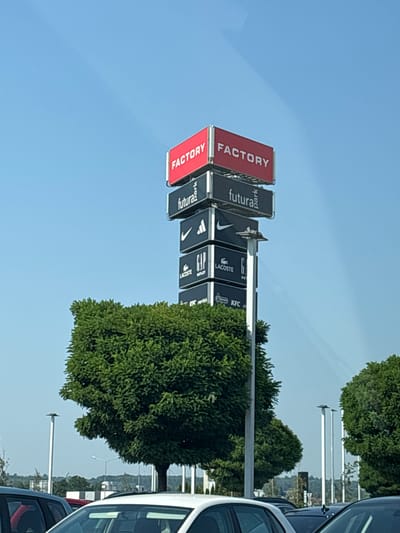The image shows a tall sign tower outside a shopping outlet in Modlniczka, Poland. The tower has multiple signs stacked vertically, with the top sign reading "FACTORY". Below it are signs for "futura park", "NIKE", "GAP OUTLET", "LACOSTE", and "KFC". The sign tower is located in an open area with blue sky above and some green trees and cars in the foreground. The scene is captured during the daytime, with clear weather conditions.
