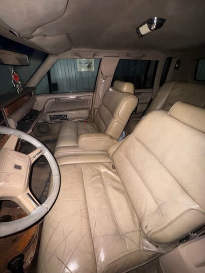 The image displays the interior of an unoccupied light brown/beige vehicle, identified as being in Zhabkino, Russia, based on external cues. The perspective is from the driver's side floor, looking across the front passenger area towards the rear seats.
**Subjects and Objects:**
The primary subject is the vehicle's interior. A beige four-spoke steering wheel, showing wear on its rim, is partially visible in the lower left foreground. The seating consists of worn, tan leather or faux leather upholstery, exhibiting numerous creases, cracks, and some discoloration across all visible seats. The front passenger seat is reclined. A dashboard with a dark upper surface and a prominent wooden trim accent is visible on the passenger side. A black rose-shaped air freshener hangs from the rearview mirror, which is partially visible. An overhead dome light fixture with a chrome base is mounted on the vehicle's ceiling. Door panels, matching the interior color, are visible, featuring power window controls.
**Environment:**
Through the vehicle's side and rear windows, a light grey-green corrugated metal wall is visible, indicating the vehicle is parked inside a garage, storage unit, or similar enclosed structure. A white piece of paper with dark Cyrillic text is taped to this external wall, visible through the front passenger window, confirming a Russian setting. The interior is dimly lit, suggesting low ambient light conditions.
**People, Actions, and Interactions:**
No people are present in the image. No actions or interactions are depicted. The scene is static.
