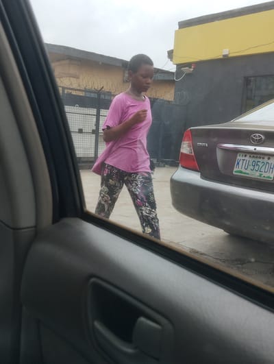 The image shows a young person walking in front of a dark-colored Toyota sedan parked on a street. The person is wearing a pink t-shirt and patterned pants. The car has a Lagos license plate. In the background, there is a building with a yellow and grey facade, and a metal gate with barbed wire. The scene appears to be in Alagbede, Nigeria, as indicated by the license plate. The sky is overcast.