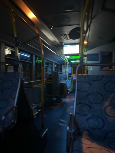 The interior of a bus is captured in a blurry image. Rows of seats with blue patterned upholstery stretch down the aisle. Yellow handrails run along the ceiling. A digital display screen glows near the front. Windows line the sides, and luggage racks are visible above the seats. The overall lighting is dim.