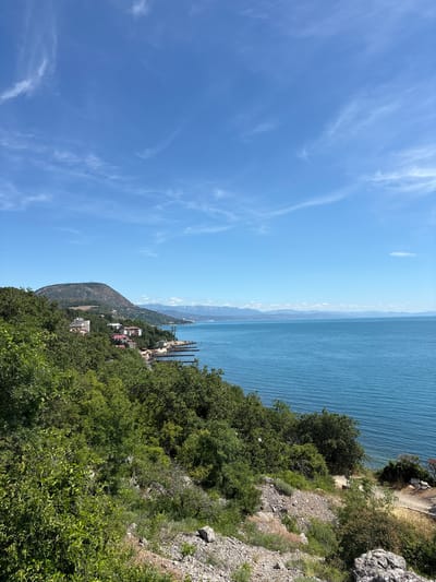 This image depicts a coastal scene with a clear blue sky and calm sea. In the foreground, a rocky, vegetated slope descends towards the water. The middle ground features a town with white buildings, some with red roofs, nestled along the coastline, surrounded by lush green trees. Beyond the town, a prominent, tree-covered hill rises, topped with what appear to be communication towers or antennas. In the distance, a range of hazy blue mountains forms the horizon. The sea stretches out to the right, meeting the distant mountains under a sky dotted with wispy white clouds. The location is Bondarenkovo, Ukraine.