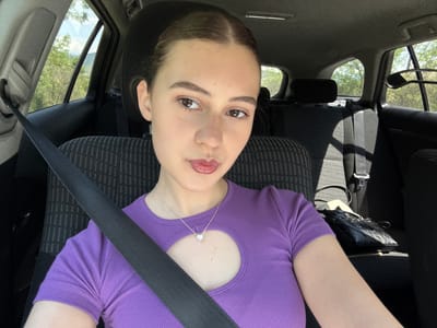 A fair-skinned young woman with brown hair pulled back is shown from an upside-down perspective in the interior of a car. She is wearing a purple, ribbed shirt with a circular cutout below the neck and a silver necklace with a small pendant. She is wearing a black seatbelt diagonally across her torso. The car's seat is upholstered in a patterned fabric. The interior of the car's roof, windows, headrest, and dashboard are visible. Through the car window, trees and sunlight can be seen. The city of Levski, Bulgaria is not identifiable from the image.