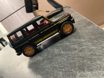 A black toy Mercedes-Benz G-Class is positioned on a textured gray surface. The toy car features gold accents, including stripes along the body and a gold emblem on the wheel. The interior appears to be red. The background shows a blurred office-like setting with a black monitor and a light-colored desk or shelf. No people are present in the image. The scene is indoors. The location of Petrovsk-Zabaykalskiy, Russia, is not evident from the image.