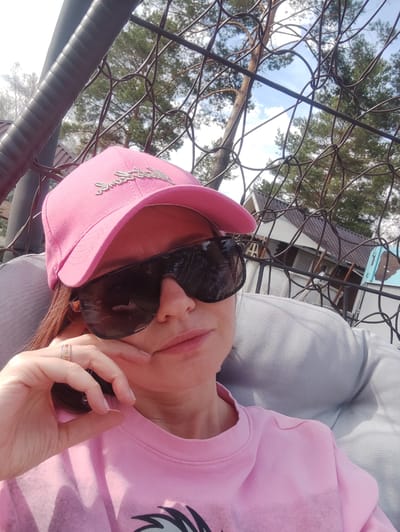 A woman is sitting in a wire-framed chair outdoors, wearing a pink baseball cap with silver lettering, dark sunglasses, and a pink t-shirt. She is resting her cheek on her hand, which has a ring on one of the fingers. The background shows trees and a building, suggesting a setting in the town of Baklashi, Russia.