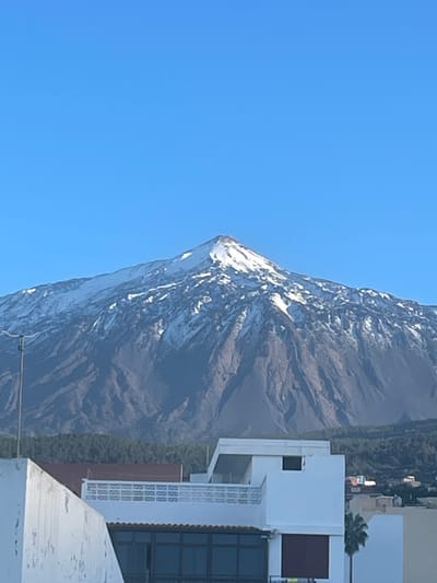 A towering, snow-capped mountain dominates the frame, its peak sharply defined against a clear blue sky. The mountain's slopes are rugged and textured. To the right, a modern building with glass and white walls stands near a green area with trees. The composition juxtaposes natural grandeur with human architecture.
