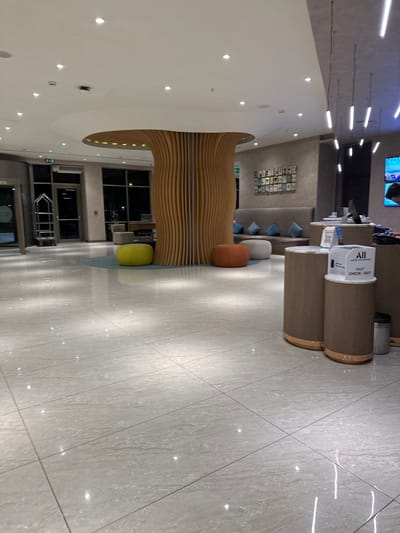 This image depicts the brightly lit, modern lobby of the Novotel Istanbul Zeytinburnu hotel, located in Zeytinburnu, Turkey.

The scene features a polished, light-colored tiled floor with a reflective surface, illuminated by numerous recessed spotlights in a multi-tiered white ceiling. A prominent central feature is a large, mushroom-shaped decorative structure composed of vertical wooden slats forming the base, topped by a flat, white cap with integrated lighting along its underside. Surrounding the base of this structure is a circular light blue rug, on which three rounded, upholstered ottomans are placed: one lime green, one orange, and one light grey.

To the left, a glass revolving door serves as an entrance, with a silver, two-tiered luggage trolley positioned nearby. Beyond this, additional glass doors and dark windows indicate the exterior is dark, suggesting evening or night.

On the right side of the image, a reception or check-out area is visible, featuring multiple cylindrical structures clad in a brown material with copper-colored base rings. One of these structures holds a white box labeled "ALL FAST CHECK-OUT." A computer monitor is also present on this counter. Further right, a large blue screen, likely a digital display, shows "NOVOTEL" branding and a partial image of a person.

In the background, a long, grey upholstered bench or sofa with light blue throw pillows is positioned against a textured beige wall adorned with a grid of framed pictures or art. The area is devoid of people, actions, or direct interactions. The environment is well-maintained and appears unoccupied.