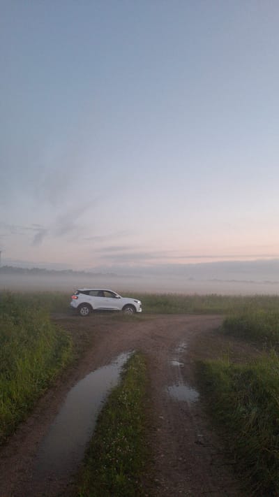 A white SUV is parked on a dirt road in a field near Kaporskoe, Russia. The sky is a soft gradient of blue and pink, suggesting either sunrise or sunset. A light fog hangs over the distant tree line, softening the horizon. The road itself is muddy with puddles, indicating recent rain. Tall green grass borders the road on both sides. The overall atmosphere is tranquil and slightly ethereal. There are no people visible in the image.