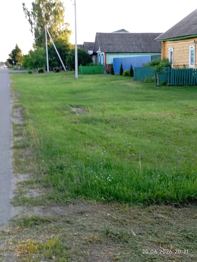 The image depicts a rural scene in Урыцкі сельскі Савет, Belarus. In the foreground, a paved road runs along the left side of the frame, bordered by a strip of grass and dirt. The middle ground is dominated by a wide expanse of green grass, with some areas appearing freshly cut or disturbed. Beyond the grassy area, a series of houses and fences are visible. To the right, a yellow wooden house with white-trimmed windows stands. Adjacent to it is a green wooden fence. Further left, a blue metal fence is present, behind which a long, grey-roofed building is situated. Several green fences are scattered throughout the scene. In the background, trees are visible, some of which are tall and leafy. The sky is overcast and bright. A timestamp in the bottom right corner reads "20.06.2026 20:21". No people are clearly visible in the image.