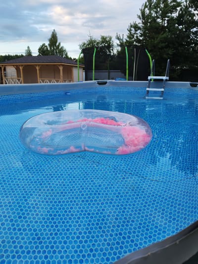 The image shows an inflatable pool float with pink filling floating in a blue swimming pool. The pool is tiled with a hexagonal pattern. In the background, there is a wooden gazebo with a dark roof and a trampoline with green poles and a ladder. Trees and a cloudy sky are visible beyond the pool. The scene appears to be set in a backyard on a summer day, possibly in the late afternoon or early evening given the lighting. The location is identified as Kstovo, Russia. No people are visible in the image, and there is no visible text.