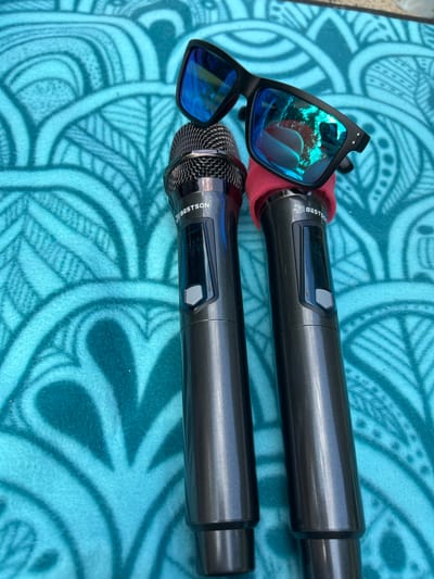 Two dark gray microphones are placed on a turquoise towel with a circular pattern. One microphone has a red foam windscreen on its head. A pair of black sunglasses with blue mirrored lenses rests on top of the microphones. The sunglasses reflect a person holding a camera. The scene is outdoors, as indicated by the bright lighting and reflections. The exact location is not specified in the image or its context.