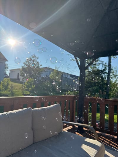 This image depicts a sunny outdoor scene, likely a backyard deck, with numerous bubbles floating in the air. The foreground features a portion of an outdoor sofa with beige cushions, casting a shadow from sunlight filtering through an overhead structure. A wooden railing is visible behind the sofa, and beyond that, a green lawn and trees are seen. In the background, houses and more trees are visible under a clear blue sky. The sun is prominent in the upper left quadrant, with lens flare and light rays extending outwards. The bubbles are scattered throughout the frame, some appearing iridescent. There are no people or animals visible in the image. The location in Dieppe, Canada, is not explicitly indicated in the image itself.