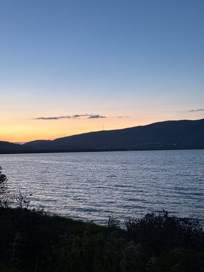 The image depicts a twilight scene over Lake Sevan, with the city of Tsamakaberd, Armenia, faintly visible in the distance along the base of a mountain range. The sky transitions from a deep blue at the top to a soft orange and yellow gradient near the horizon, indicating sunset or sunrise. A few wispy clouds are scattered across the sky. The foreground features a dark, silhouetted landscape of foliage and trees on a gentle slope leading down to the water. The surface of Lake Sevan is calm, with gentle ripples reflecting the subdued light. The distant shoreline is marked by a dark line, with scattered lights of Tsamakaberd illuminating the foothills of the mountains. A tall, slender tower, likely a communication tower, stands prominently on the crest of one of the mountains. No people or animals are visible in the frame.