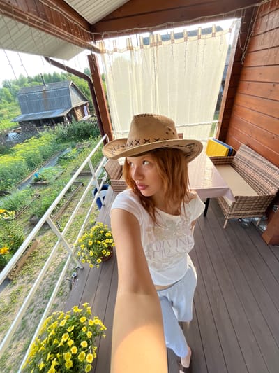 A young woman with reddish hair, wearing a light-colored t-shirt with a graphic design and light blue sweatpants, and a brown cowboy hat, stands on a wooden porch, taking a selfie. The porch has a white railing and is decorated with potted yellow flowers. In the background, a rural landscape is visible, with a small weathered house, a garden with raised beds, and lush green vegetation. The porch is partially covered by a wooden roof with string lights. To the right of the woman, a table covered with a pink tablecloth and a wicker bench are visible. The scene appears to be set in a rural area outside of Суроватиха, Russia, given the architectural style and the surrounding vegetation.