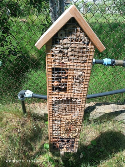 In a garden environment, a wooden insect hotel stands vertically. The hotel is constructed from natural materials such as hollow reeds, wood, and pine cones, and is covered by a wire mesh. A black hose with a blue connector runs horizontally behind the insect hotel. The ground is covered with grass, and in the background, a wire fence is visible. The image was captured on July 6, 2020, at 12:16 PM. The specific location is Vita-Poshtova, Ukraine.