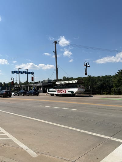 The image shows a street scene in Wisconsin Dells, Wisconsin, United States of America. In the foreground, there is a road with two lanes of traffic and a concrete sidewalk. On the right side of the road, there is a green bike lane. In the middle ground, there is a duck boat, which is a tour vehicle that can travel on both land and water. The duck boat is white with a green and white striped awning and has "DUCKS" written on its side. Behind the duck boat, there is a building with a sign that says "THE DISTRICT." There are several cars parked along the side of the road. In the background, there are trees and a blue sky with some clouds. The scene appears to be a sunny day.