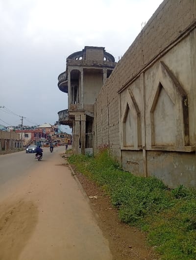 This image captures a daytime street scene at Itakpe Junction, Nigeria, under an overcast sky. In the immediate foreground to the right, a narrow strip of unkempt vegetation and bare earth parallels a paved road. Adjacent to this strip, a long concrete block wall, partially rendered, extends into the background, featuring three recessed, pointed arch decorative elements along its length.

Dominating the midground is a large, multi-story building, appearing unfinished or under construction, composed of concrete and blocks. This structure features a prominent circular upper section with multiple supporting columns and arched openings, alongside several balconies with visible railings on lower levels. Exposed rebar protrudes from the top edge of the concrete block wall immediately next to this building, indicating ongoing construction.

The paved road occupies the central and left portions of the image. Several vehicles are observed traveling away from the viewer: a dark-colored sedan with activated headlights, followed closely by an individual on a dark motorcycle wearing a dark jacket. Further down the road, another person on a dark motorcycle and a partially visible three-wheeled auto-rickshaw are also in motion. On the left side of the road in the background, a pedestrian in dark clothing is observed walking.

The left side of the street is lined with various low-rise buildings, many appearing residential or commercial, some seemingly unfinished or in disrepair. Overhead power lines and utility poles are visible along this side of the road.