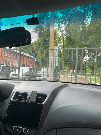 This media file shows the interior of a car looking out through the windshield. The scene outside appears to be a street with a metal fence, trees, and buildings. There are several objects visible inside the car, including a dashcam mounted on the windshield and a black screen which could be a car stereo or navigation system. An air freshener is clipped to an air vent.

The sunlight suggests it is daytime, likely overcast given the muted light. The presence of trees and buildings suggests an urban or suburban environment. The license plates visible on the cars outside are in Cyrillic script, indicating a Russian context, consistent with the provided location of Monastyrshchina, Russia. The overall mood is neutral, with no discernible emotions. The visible text on the air freshener reads "areon".