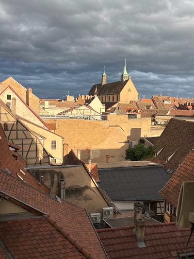 The image depicts a rooftop view of the historic city of Colmar, France, with a dramatic sky overhead. The architecture suggests the traditional half-timbered style common in Alsace, with steeply pitched terracotta tiled roofs. In the background, a church with a green steeple stands prominently, a common landmark in European cities. The lighting indicates it is either early morning or late afternoon, with strong shadows cast across the buildings. The presence of air conditioning units on some rooftops suggests modern additions to the older structures. The scene is devoid of people, focusing solely on the urban landscape and the interplay of light and shadow under a cloudy sky.