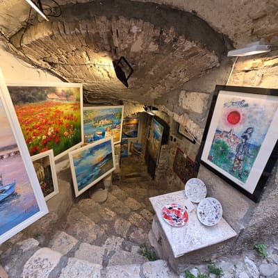 This is a medium-shot, eye-level view of an art gallery located in Saint-Paul-de-Vence, France. The gallery is set within a rustic, stone-arched space with uneven, natural stone steps leading downwards. The lighting is dim, with several modern, track-mounted LED lights illuminating the artworks.

The walls and ceiling are made of rough, uneven stone and brick, with exposed electrical wiring visible. Various paintings and decorative plates are displayed on the steps and walls.

Prominent artworks include:
- A large painting of a field of red poppies in the foreground, with houses in the background.
- Several seascape paintings featuring coastal towns and boats.
- A framed print of a colorful, surreal artwork depicting figures and a red sun over a town, with what appears to be a signature at the bottom right.
- Three decorative plates are displayed on a stone pedestal, one with a vibrant abstract design and two with intricate patterns.
- Other smaller paintings and framed art are visible further down the steps and on the walls.

The overall impression is of a charming, somewhat dimly lit art space that highlights the artistic creations within its historic, natural stone architecture.