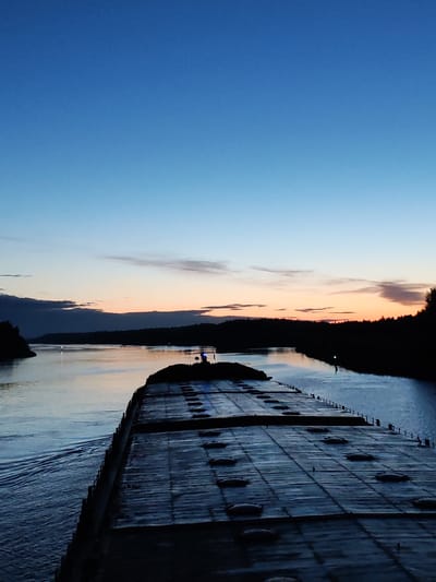 The image shows a view from the deck of a barge or large vessel navigating a body of water, likely a canal or wide river, at sunset or dusk. The sky is transitioning from blue to orange near the horizon, with some scattered clouds. The banks of the waterway are visible as dark, silhouetted land masses with trees. The deck of the barge is in the foreground, showing a pattern of what appears to be cargo covers. The location of Badozhskoi Pogost, Russia, is not explicitly confirmed in the image, but based on the landscape and type of vessel, it could be consistent with waterways found in that region.