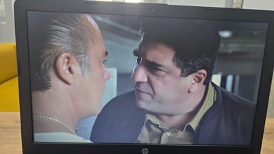 The image shows a close-up of two men on a laptop screen. The man on the left has slicked-back grey hair and is wearing a gold chain. The man on the right has dark, curly hair and is wearing a greenish-brown collared shirt and a dark jacket. They appear to be in a serious conversation. The laptop is on a wooden desk, with a yellow object partially visible in the background. The scene is viewed from an angle, with the laptop screen taking up most of the frame. No specific location in Oral, Kazakhstan is depicted.