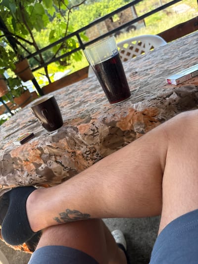The image presents a close-up, angled view of a person's lower body and a patterned table in an outdoor or semi-outdoor setting. The perspective is from above, looking down.
In the center and right, the bare, slightly hairy legs of an individual wearing blue shorts are visible. On the lower part of the right leg, above a black ankle sock, there is a dark tattoo featuring an abstract design or figure with some illegible script underneath. The left foot is also wearing a black ankle sock.
The table surface, which occupies the left and central areas, has a distinct mottled pattern resembling marble or granite, with shades of beige, brown, and black. On the table, a clear glass filled with a dark red beverage, possibly wine or a dark soda, is prominently featured. The glass has some condensation and a small, partially visible blue and white label or logo near its rim. Nearby, a dark brown ceramic mug with a handle is placed. Also on the table are two small rectangular items: a matchbox with a red and yellow design, and a white and pink cigarette pack. The cigarette pack clearly displays a health warning: "WARNING: SMOKING KILLS" along with other faint text, likely including the brand name "Rothmans."
The background reveals a vibrant green natural environment, with lush foliage, possibly vines or dense leaves, seen through a dark metal railing. A white plastic garden chair is partially visible behind the table on the left side. The scene is brightly illuminated by natural daylight, suggesting a sunny day. The overall impression is one of leisure and relaxation in a natural, possibly rustic, environment, consistent with a rural location such as Staro Selo, Bulgaria. The activity depicted appears to be casually sitting outdoors, enjoying a drink and possibly smoking.
