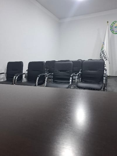 The image depicts an unoccupied meeting room located in Kumaryk, Uzbekistan. The foreground is occupied by a dark, reflective rectangular table surface. Behind the table, multiple rows of identical black office chairs with chrome frames and armrests are uniformly arranged, facing away from the camera. The room features plain white walls and a white ceiling equipped with overhead artificial lighting, which casts reflections on the tabletop. In the background, positioned in the right corner of the room, a white flag hangs vertically. The flag displays an emblem near its top and vertical text in both Cyrillic and Latin scripts, including the words "SPORT" and "VAZIRLIGI," consistent with "O'ZBEKISTON RESPUBLIKASI SPORT VAZIRLIGI" (Ministry of Sport of the Republic of Uzbekistan). No individuals are present, and no actions or interactions are occurring within the visible scene.