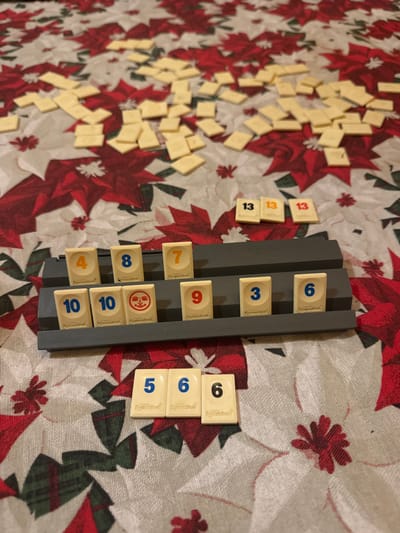 A Rummikub game is in progress. Several tiles are scattered on a tablecloth with a poinsettia pattern. A dark gray tile holder displays the following numbers: 4, 10, 10, 8, a joker, 7, 9, 3, and 6. Three number 13 tiles sit next to the holder. To the right are tiles showing a set of 5, 6, and two 6s. The scattered tiles are likely the remaining pieces from which players draw.