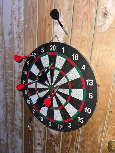 The image displays a dartboard mounted on a textured, light brown wooden planked surface, likely a wall or ceiling, viewed from a slightly upward angle. The planks are horizontally oriented and show signs of wear, including some lighter, possibly painted or discolored, areas. In the upper right corner, a small, ornate metallic fixture, possibly part of a light or door mechanism, is visible.

A game of darts appears to be in progress or has recently concluded, as several darts are scattered around and within the dartboard.
- One black and silver dart is stuck in the wooden plank surface to the immediate left of the dartboard, its tip embedded in the wood.
- Two black darts with red flights are embedded close together in the central bullseye region of the dartboard.
- Two other black darts with red flights have fallen out of the dartboard and are resting on the wooden plank surface directly below the board, pointing downwards. One is positioned below the '14' segment, and the other below the '5' segment.
- Two silver and black darts are embedded in the lower right section of the dartboard, one in the single '8' segment and another in the single '16' segment.

The dartboard itself is round, featuring the standard scoring layout with black and white segments, and red and green rings for double and triple scores. The numbers 1 through 20 are clearly visible around the outer edge of the board. In this particular orientation, the '20' segment is located at the bottom of the board. The dartboard shows significant signs of use, with numerous small puncture marks from previous throws.

The lighting is bright, casting some shadows from the darts. The overall setting suggests a casual, indoor recreational area.
