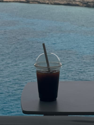 A clear plastic cup containing a dark, iced beverage is centrally positioned on a flat, light grey surface, likely a table or ledge, with a rounded corner visible on the left. The cup is fitted with a transparent dome lid and contains a dark, slender straw extending vertically into the drink. Visible condensation covers the exterior of the cup, indicating a cold beverage. The background features a vast expanse of blue ocean with subtle surface ripples, extending to a distant, low-lying landmass or shallow reef area appearing as an indistinct horizontal line. This scene is situated in the Berry Islands, Bahamas. No individuals or active interactions are present in the image.