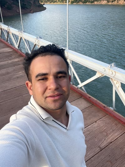 The image shows a man in a white shirt standing on a wooden bridge, likely in a tourist area. The bridge has white steel supports. The background includes water, possibly a lake or river, and rocky, reddish-brown cliffs with sparse vegetation. The scene is sunny and appears to be outdoors during daylight hours. Considering the prompt, the location is likely Ait Mazigh, Morocco. The image is likely a selfie taken by the man.