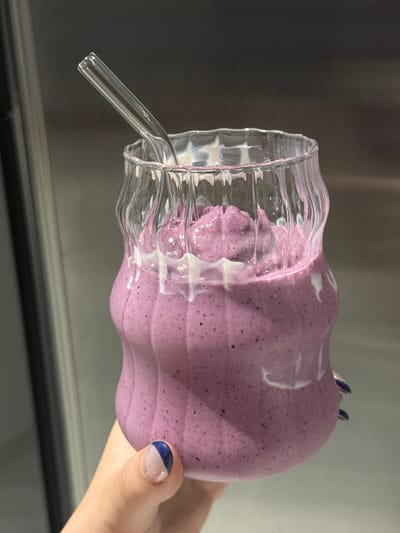 A close-up, horizontally oriented shot captures a hand holding a clear, wavy-textured glass filled with a thick, foamy, purple beverage. The drink, possibly a smoothie or milkshake, contains small dark specks, suggesting berries, and features prominent white swirls or streaks on its surface. It appears to overflow slightly over the rim of the uniquely designed glass. A clear glass straw is inserted into the beverage, pointing downwards and to the left.

The hand, positioned on the right side of the frame, has manicured nails painted with a two-toned design: a solid blue on the upper part and a clear or very light lower section.

The background is a blurred indoor setting, predominantly in shades of dark and light grey. A distinct horizontal line cuts across the lower part of the background, hinting at a countertop or a structural element. The lighting is soft and even, consistent with an indoor environment. There are no visible cues for the time of day, weather, or specific location within the image itself, beyond the general indoor setting. No text is visible. The provided context indicates the location is Zapopan, Mexico, but this is not visually represented in the media.