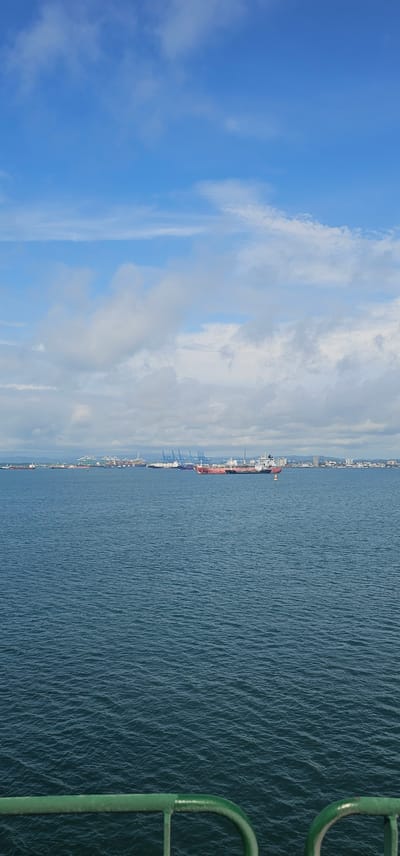 A wide-angle, vertically oriented, eye-level shot shows a bright, cloudy blue sky above a vast expanse of deep blue water. In the distance, a coastline is visible, lined with a port complex featuring numerous cranes of various colors, including blue and green, as well as a city skyline with buildings. Several large cargo ships are anchored in the water near the port. In the foreground, a curved, weathered green railing runs along the bottom of the frame, indicating the viewpoint is from a boat or pier. The water exhibits small ripples, suggesting a calm day. The overall scene depicts a busy port area in Cristóbal, Panama.
