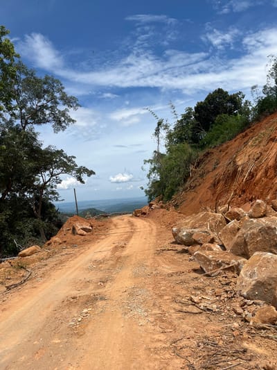 This media file captures a downward-sloping, unpaved dirt road cutting through a steep, eroded hillside in Son Dien, Vietnam. The road is made of reddish-brown soil and is uneven, with scattered rocks and debris. To the left of the road, the hillside is a sheer cut of the same reddish soil, with sparse vegetation clinging to its surface. Further down the slope, lush green trees and foliage are visible, indicating a more vegetated area below.
The sky is a vibrant blue, streaked with wispy white clouds. The lighting suggests it is daytime, likely mid-morning or afternoon, with the sun casting shadows and highlighting the textures of the earth and rocks. The atmosphere appears clear, with a distant landscape visible through the trees, hinting at a rural or mountainous setting. A single wooden post stands upright along the edge of the road, serving as a subtle marker or perhaps part of a simple structure.
There are no people or animals visible in the image. The focus is on the natural landscape and the man-made path carved into it. The scene evokes a sense of raw nature and the challenges of navigating such terrain. The image composition, with the road leading the eye downwards and into the distance, creates a feeling of depth and exploration. The overall impression is one of a remote, undeveloped area in Vietnam.
