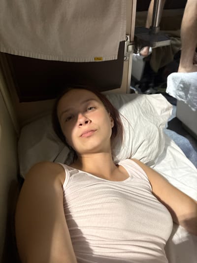 This is a horizontal, medium close-up, indoor photograph of a young woman lying on her side in bed. She is facing the camera, with her head on a pillow. Her eyes are open, and she is looking directly at the camera with a neutral expression. She has medium-length, dark brown hair that falls around her shoulders. She is wearing a white, ribbed, tank top. Her right arm is bent at the elbow and resting near her head, while her left arm is bent at the elbow and resting on her stomach. The background is out of focus, but appears to be a bedroom. There is a white pillow and white bedding. The lighting is dim, suggesting it could be nighttime or early morning. There is no visible text in the image. The overall mood of the image is relaxed and intimate.