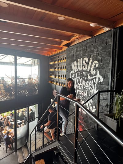 This image depicts the interior of a multi-level restaurant or bar, likely in Punta del Este, Uruguay, during the daytime. A prominent dark metal staircase with thin wire railings dominates the right side of the frame. A woman with dark hair, wearing a dark top and light-colored pants, stands on the upper landing of the staircase, holding a drink and looking towards the viewer. Below her, a man in a dark t-shirt and light shorts is visible on a lower flight of the stairs.
Behind the woman and staircase, a dark gray wall with a brick-like texture features large white painted text that reads: "MUSIC IS THE STRONGEST FORM OF..." The remainder of the phrase is not visible. Below this text, a rack filled with numerous bottles, likely wine, is integrated into the wall.
On the left side of the image, the ceiling is constructed from warm-toned wooden slats, with white circular light fixtures and a security camera mounted on it. Large windows span across the lower section of the image, revealing an outdoor dining area where many people are seated at tables enjoying meals, indicating a busy atmosphere. Reflections of the indoor space are also visible in the window panes.