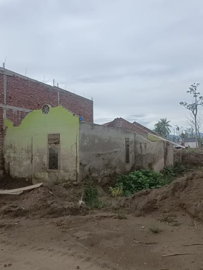 The image depicts the aftermath of damage in Gampong Blang, Indonesia, showing partially destroyed residential structures under an overcast sky. In the foreground, disturbed earth and piles of dirt suggest recent excavation or a natural disaster. Two adjoining buildings are visible, one with a red brick exterior that appears to be in the process of construction or renovation, indicated by exposed rebar along its top edge. The adjacent structure, painted in a faded lime green, has a severely damaged roofline and visible cracks in its concrete walls. One window on this building has metal bars, and the opening reveals a dark interior. Further to the right, another section of a building with a partially exposed tiled roof is seen, with more cracks and damage to the concrete walls. Lush green vegetation grows in the area between the damaged structures and the mounds of earth. In the background, several trees are visible, one appearing bare and leafless, and a small white structure. The overall scene conveys a sense of destruction and ongoing recovery efforts.