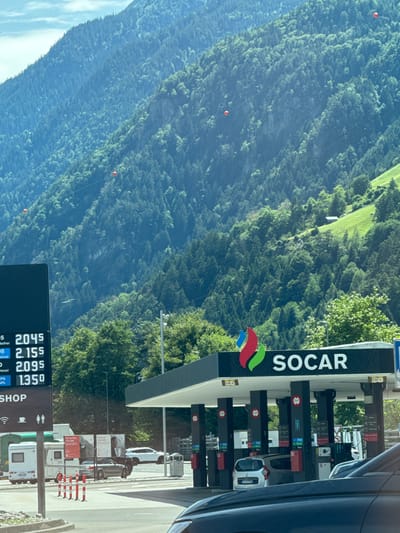 The image shows a SOCAR gas station in Schattdorf, Switzerland, set against a backdrop of a lush green, tree-covered mountain. The gas station has a modern design with a black roof and pillars. Several fuel pumps are visible, with cars parked at them. Above the pumps, the SOCAR logo is prominently displayed. A digital price board shows fuel prices, with numbers like 2.045, 2.155, 2.095, and 1.350 visible. In the foreground, the front of a dark-colored car is partially visible, indicating the image was taken from inside a vehicle. A camper van and other cars are parked in the background near the station. The mountain slope is covered in dense evergreen trees, with a small building or shed visible on a cleared section of the slope. Several red spherical objects are suspended on wires traversing the mountainside, likely part of a cableway system. The sky is a pale blue with some wispy clouds.